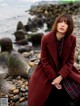 A woman in a red coat sitting on a rocky beach.